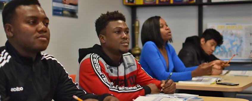 Four students take notes at their desks during a classroom lecture.