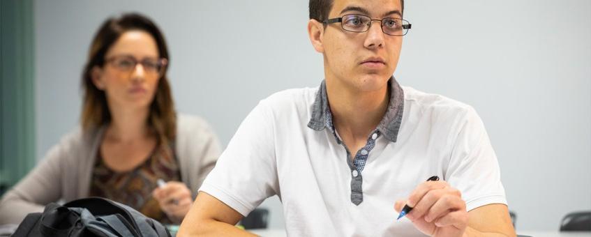 A student in a white polo and glasses holding a pencil and looking forward with another student in the background who is also looking forward.