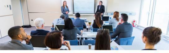 A group of employees in business casual attire sitting in a classroom with their backs to the camera while facing the two teachers standing at the front of the room on either side of a projector screen